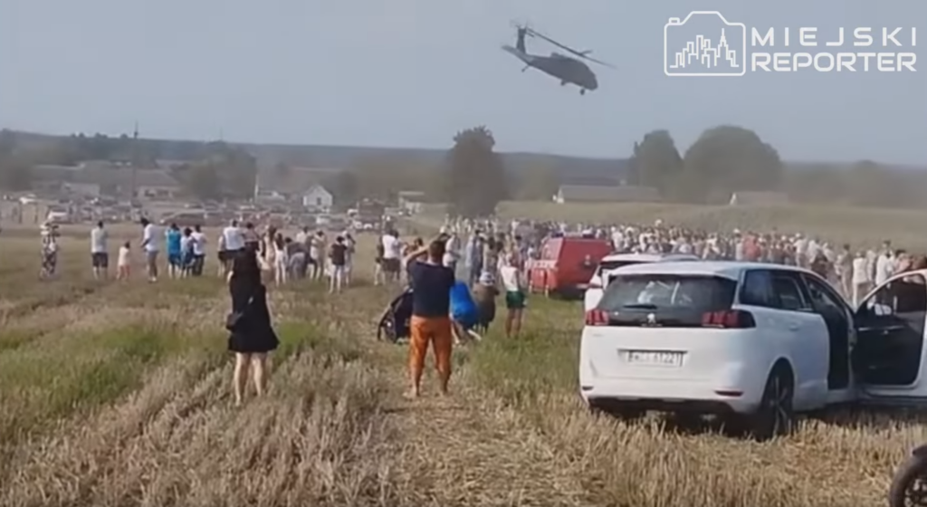 Ludzie stoją na polu, obserwując przelot śmigłowca nad zebranym tłumem i zaparkowanymi samochodami.