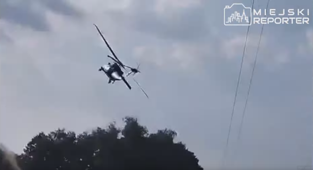 Śmigłowiec wykonujący manewr w powietrzu nad drzewami w tle i liniami energetycznymi.