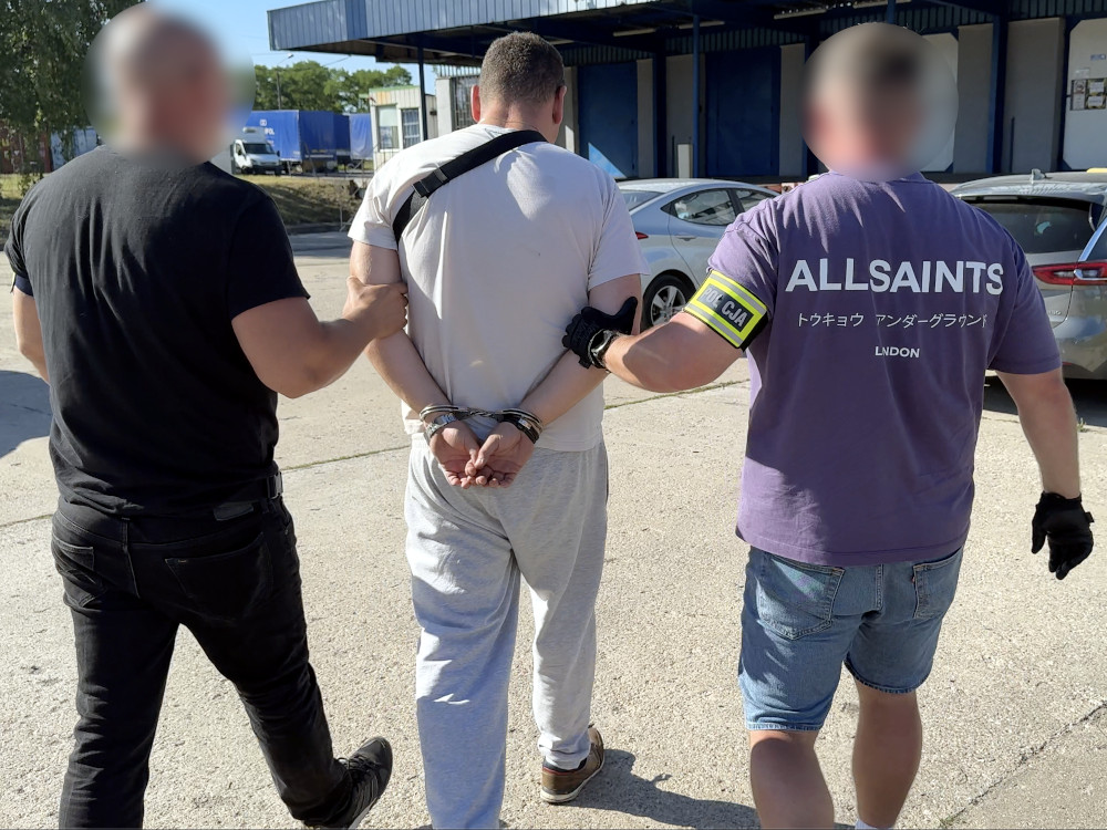 Mężczyzna w kajdankach prowadzony przez dwóch funkcjonariuszy na terenie parkingu.