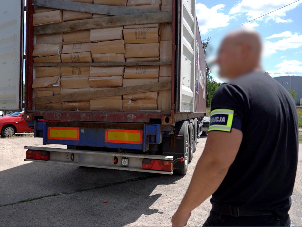 Funkcjonariusz policji obserwuje załadunek paczek w kontenerze ciężarówki na placu.