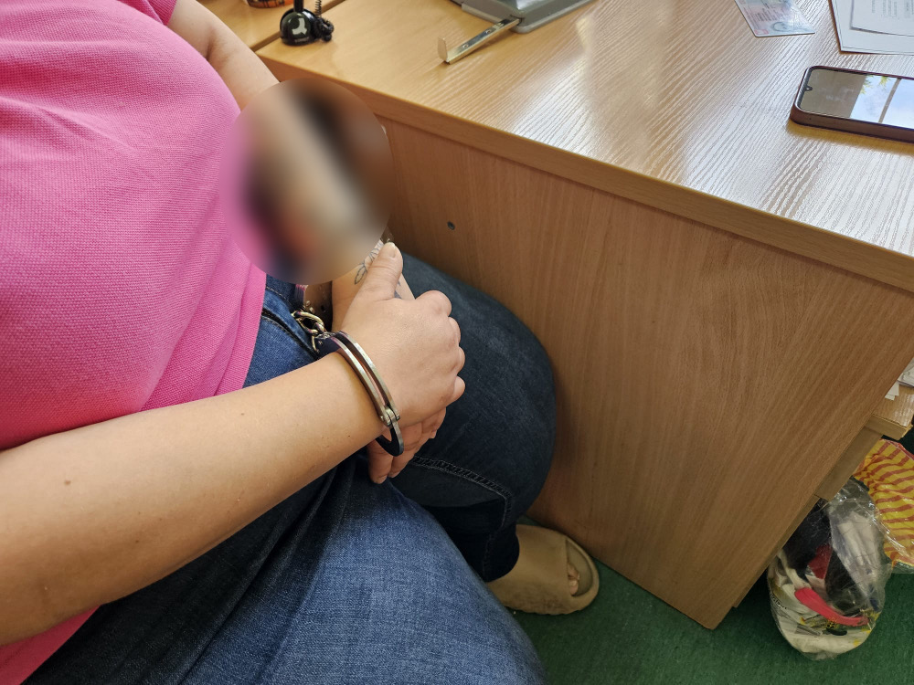 Kobieta w różowej koszulce siedzi przy biurku, z rękami związanymi w kajdankach. Na biurku znajdują się dokumenty oraz telefon komórkowy. Tło obrazu wskazuje na miejsce związane z interwencją służb. Zdjęcie ilustruje sytuację zatrzymania osoby przez organy ścigania.