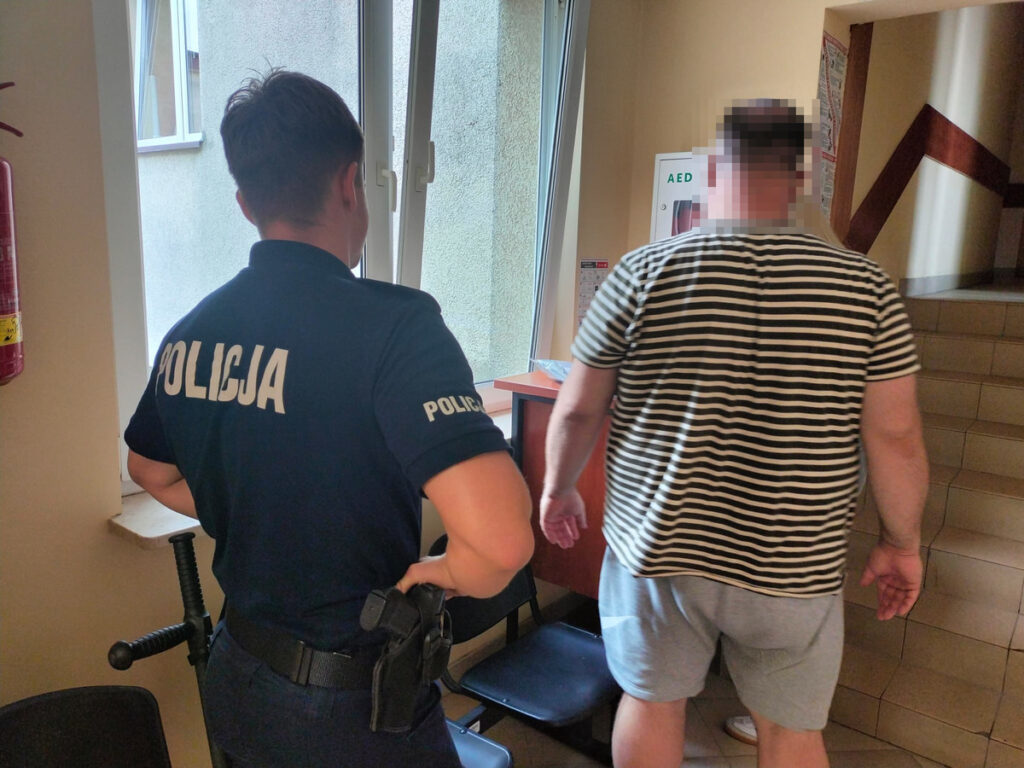 Policjant w mundurze obserwuje mężczyznę w pasiastym T-shircie idącego w stronę schodów w budynku.