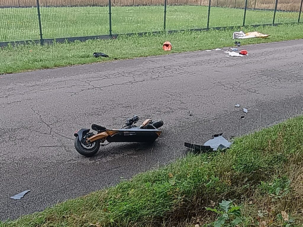 Na drodze leży przewrócony skuter elektryczny, obok rozsypane fragmenty kasku i inne przedmioty.