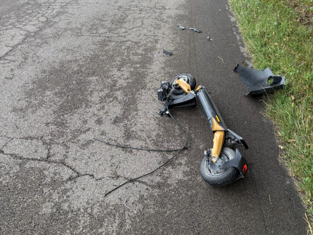 Złamana hulajnoga leży na asfaltowej drodze, obok widoczne fragmenty plastikowych osłon.