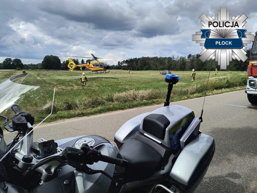 Helikopter ratunkowy ląduje na łące, a w tle widać służby ratunkowe i policyjny motocykl.