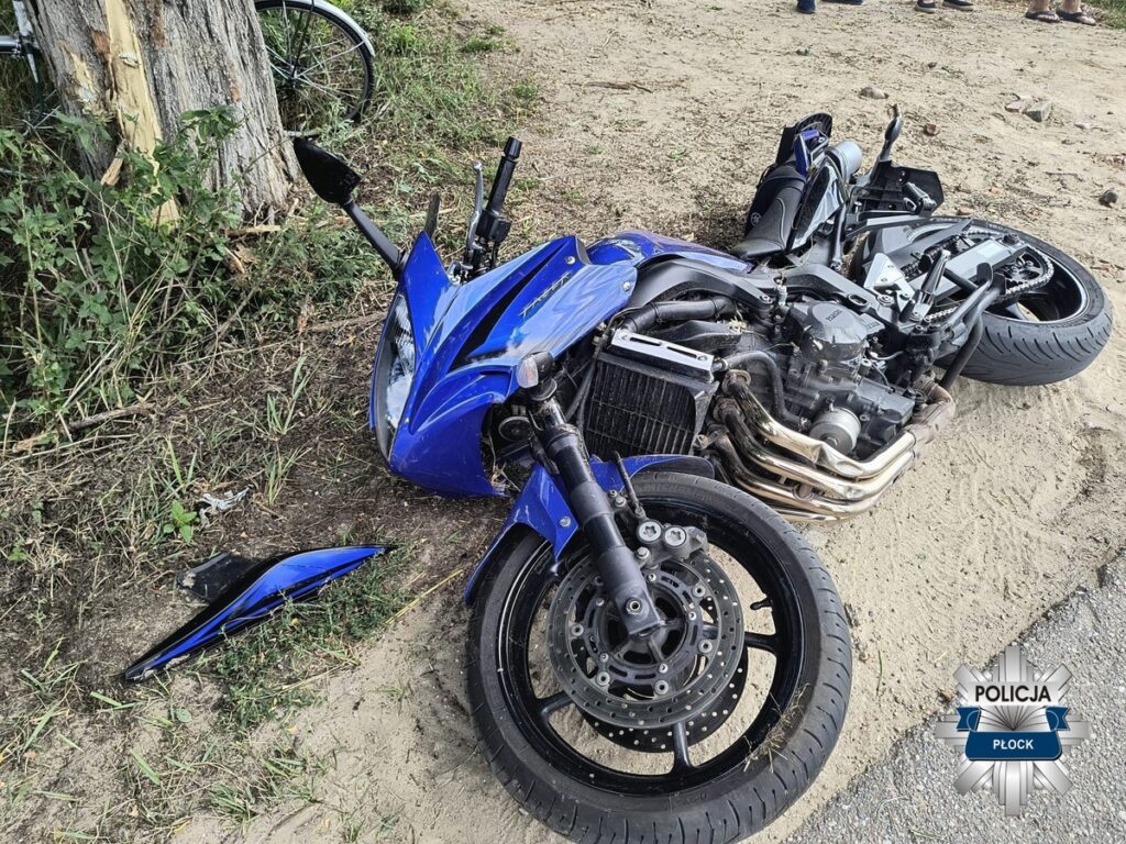 Nieprzytomny niebieski motocykl leży na boku na piaszczystym poboczu drogi, obok widoczne uszkodzone elementy.