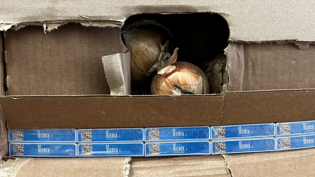W kartonie widoczne są cebule oraz paczki papierosów z niebieskimi opakowaniami.