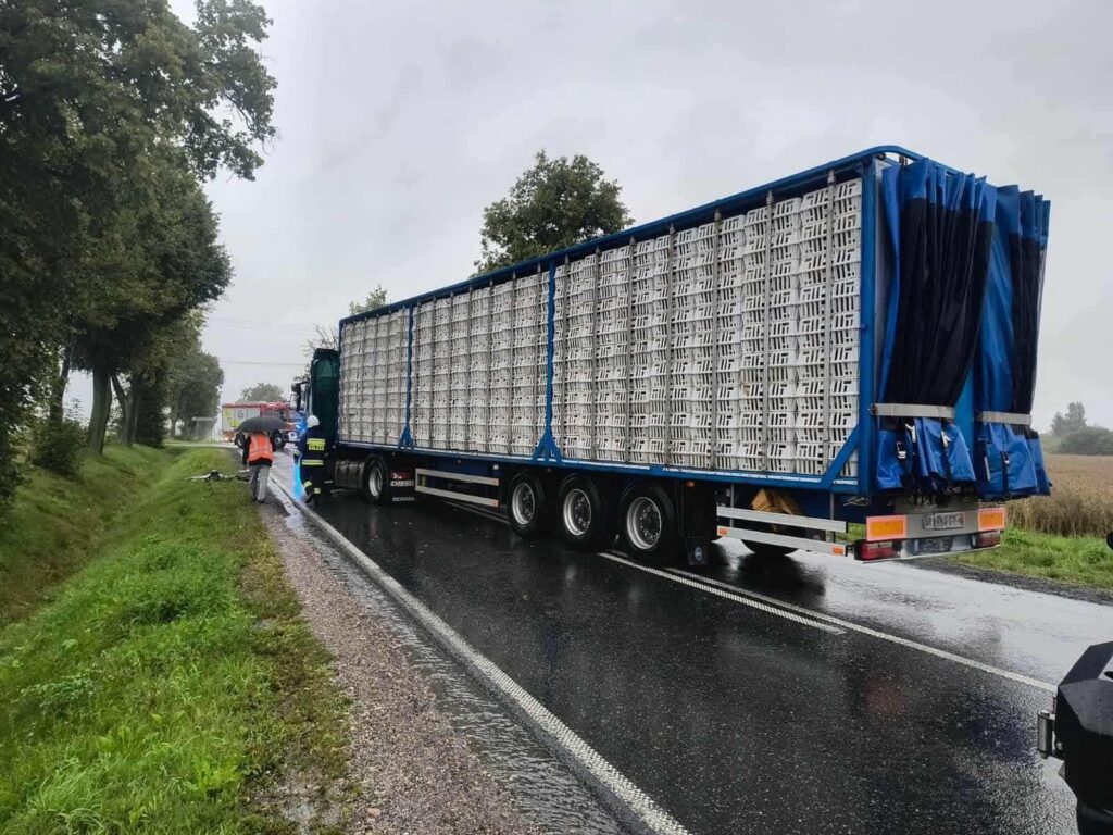 Kierowca ciężarówki rozładowuje towar na poboczu drogi w deszczową pogodę, obok pojazdu straży pożarnej.