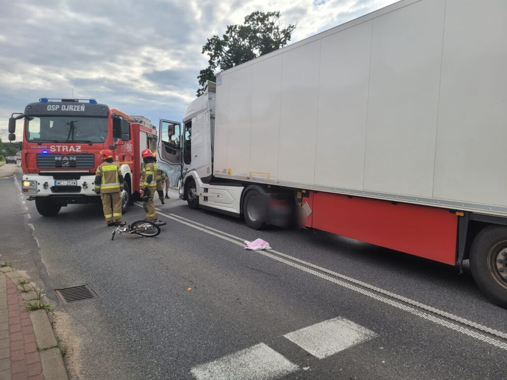 Na zdjęciu widać zdarzenie drogowe, w którym uczestniczy ciężarówka oraz pojazd straży pożarnej. Strażacy udzielają pomocy na miejscu wypadku, obok leży rower. W tle widoczna jest ulica. Sytuacja wymaga interwencji służb ratunkowych.