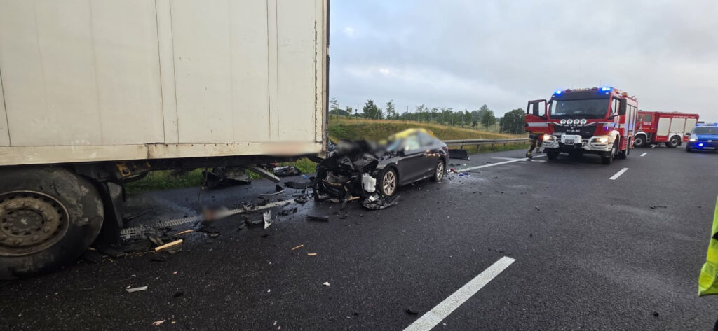 Wypadek drogowy z udziałem osobowego samochodu i ciężarówki na autostradzie, w tle widać pojazdy służb ratunkowych.