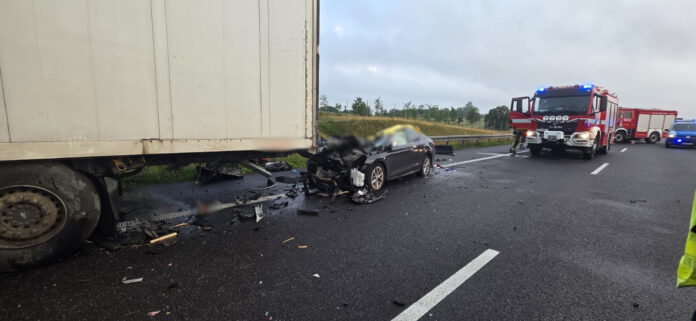 Wypadek drogowy z udziałem osobowego samochodu i ciężarówki na autostradzie, w tle widać pojazdy służb ratunkowych.