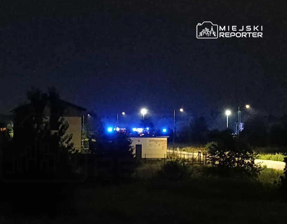Nocna scena z niebieskimi światłami pojazdów służb ratunkowych w tle, w otoczeniu budynków i ulicy.