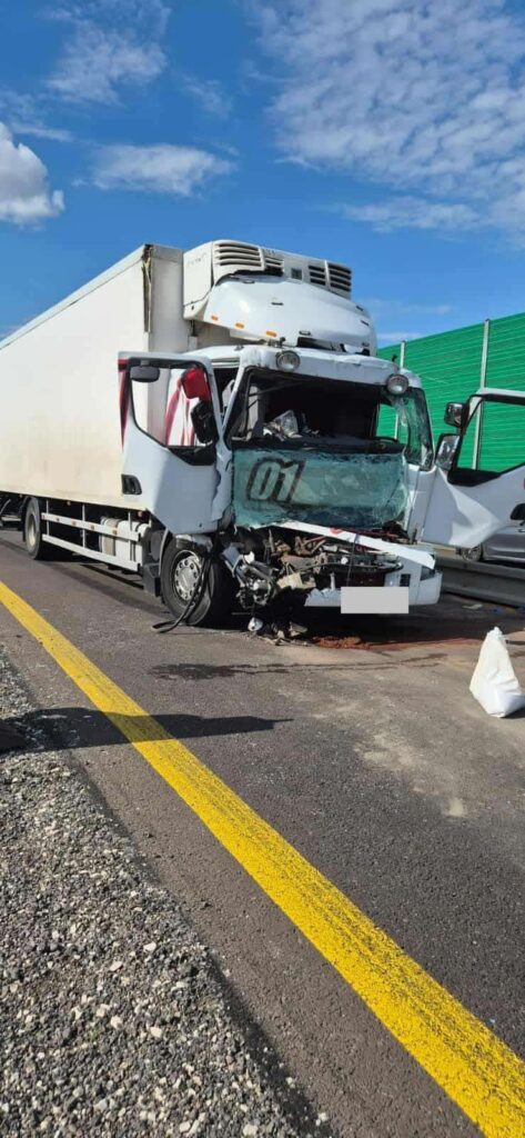 Zniszczona ciężarówka z wybitym przednim szybą stoi na drodze, obok widoczne rozlane płyny.