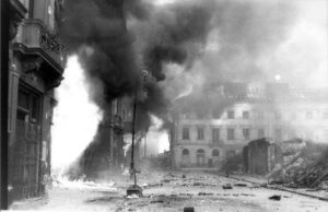 Ulica pokryta gruzem, z płomieniami i gęstym dymem unoszącym się z budynków w tle.