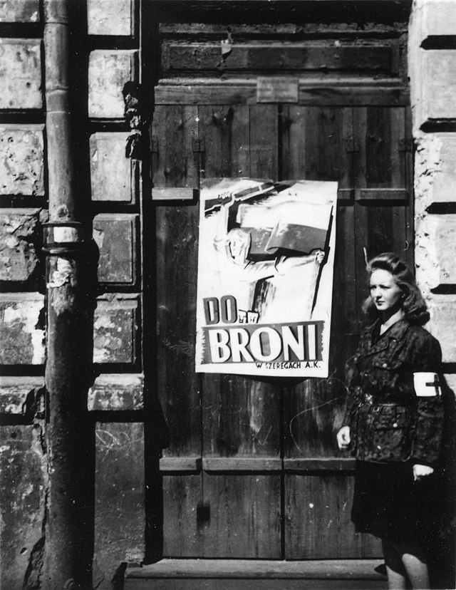 Kobieta w mundurze z opaską na ramieniu stoi obok drzwi z plakatem "DO BRONI" w tle.