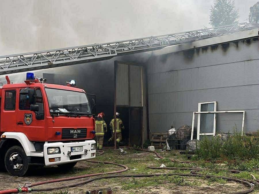Czerwony wóz strażacki z niebieskimi światłami stoi przed budynkiem, z którego wydobywa się dym. Strażacy w mundurach przystępują do akcji gaśniczej.