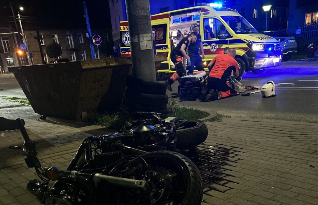 Na jezdni leży przewrócony motocykl, obok ratownicy medyczni udzielają pomocy poszkodowanemu, a w tle widać ambulans.