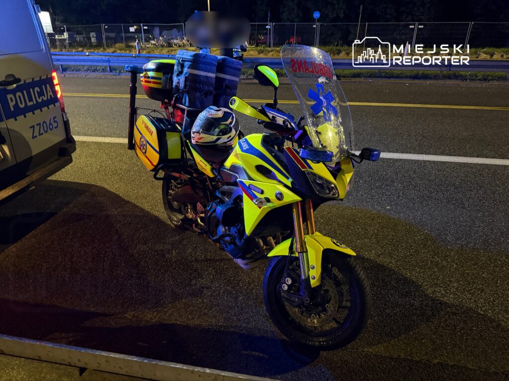 Motocykl medyczny w jaskrawym kolorze stoi na drodze obok policyjnego pojazdu, w tle widać oświetlone miasto.