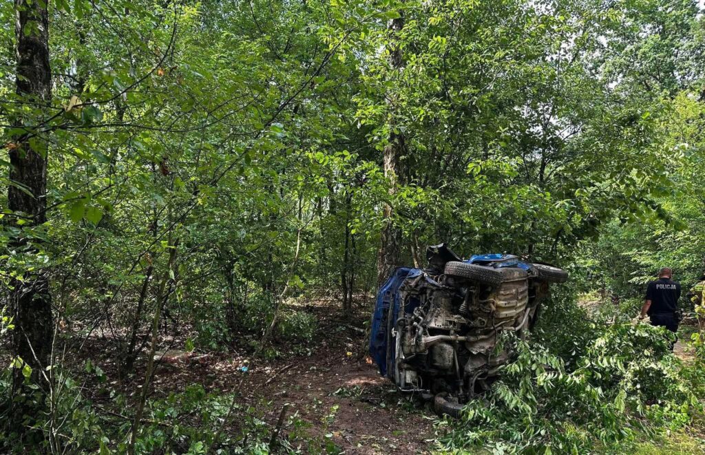 W lesie znajduje się przewrócony niebieski samochód osobowy, otoczony gęstą roślinnością. W tle widoczny jest funkcjonariusz policji, który prowadzi działania na miejscu zdarzenia. Pojazd wygląda na uszkodzony, a teren wokół jest zanieczyszczony gałęziami i liśćmi.