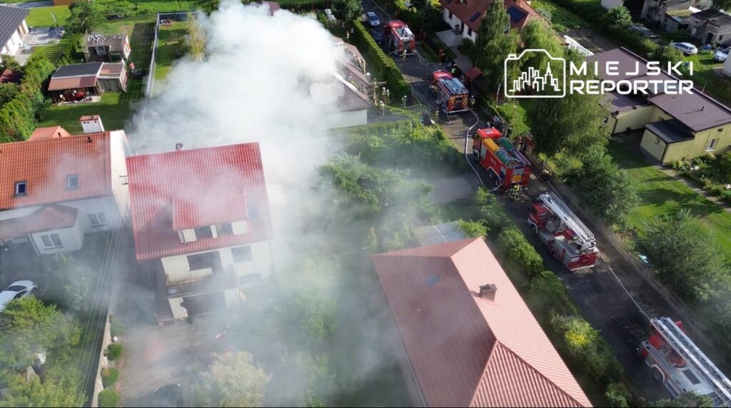 Pożar w zabudowie jednorodzinnej w miejscowości X. Na miejscu interweniują jednostki straży pożarnej, które prowadzą akcję gaśniczą. W tle widoczne są pojazdy strażackie oraz gęsty dym unoszący się nad budynkami. Mieszkańcy są ewakuowani dla ich bezpieczeństwa.