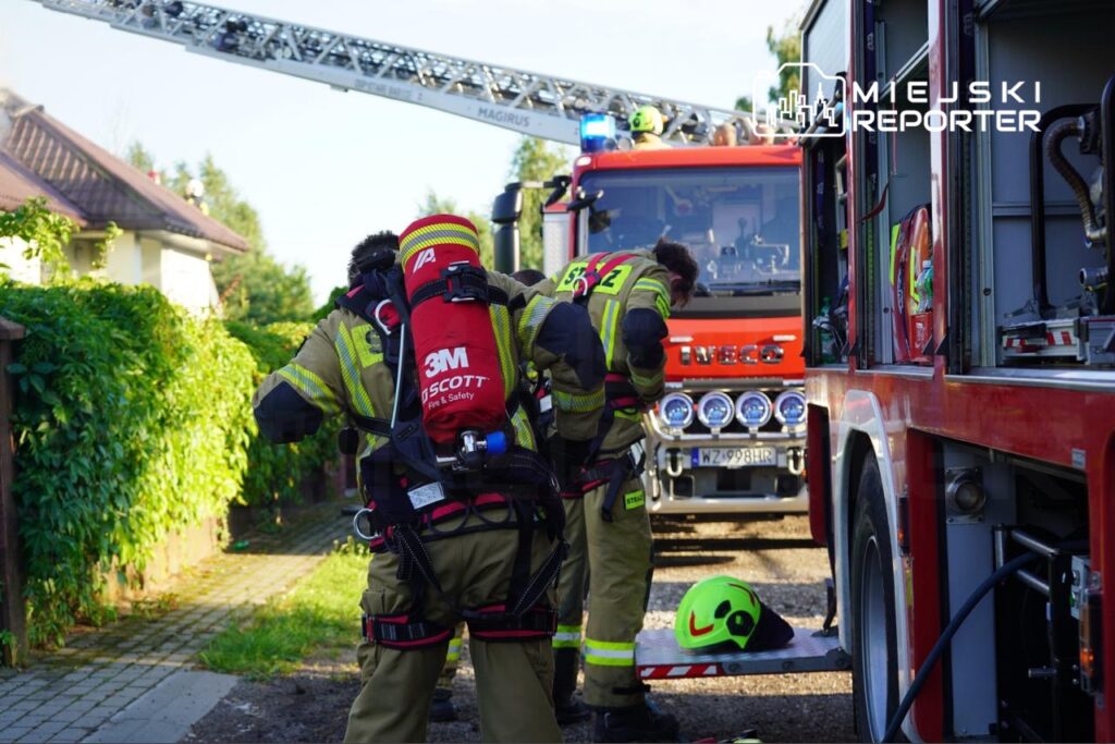 Strażacy w pełnym umundurowaniu przygotowują się do akcji gaśniczej przed wozem strażackim. W tle widoczna jest drabina strażacka oraz pojazd marki Iveco. Na miejscu zdarzenia panuje intensywna atmosfera, a strażacy sprawdzają sprzęt przed interwencją.