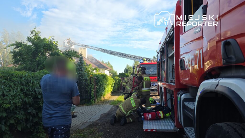 Na zdjęciu widoczna jest akcja ratunkowa prowadzona przez strażaków, którzy interweniują przy pożarze budynku mieszkalnego. W tle widać wóz strażacki oraz drabinę, na której znajdują się ratownicy. Mieszkańcy obserwują działania służb ratunkowych. Zdarzenie miało miejsce w miejscowości X.