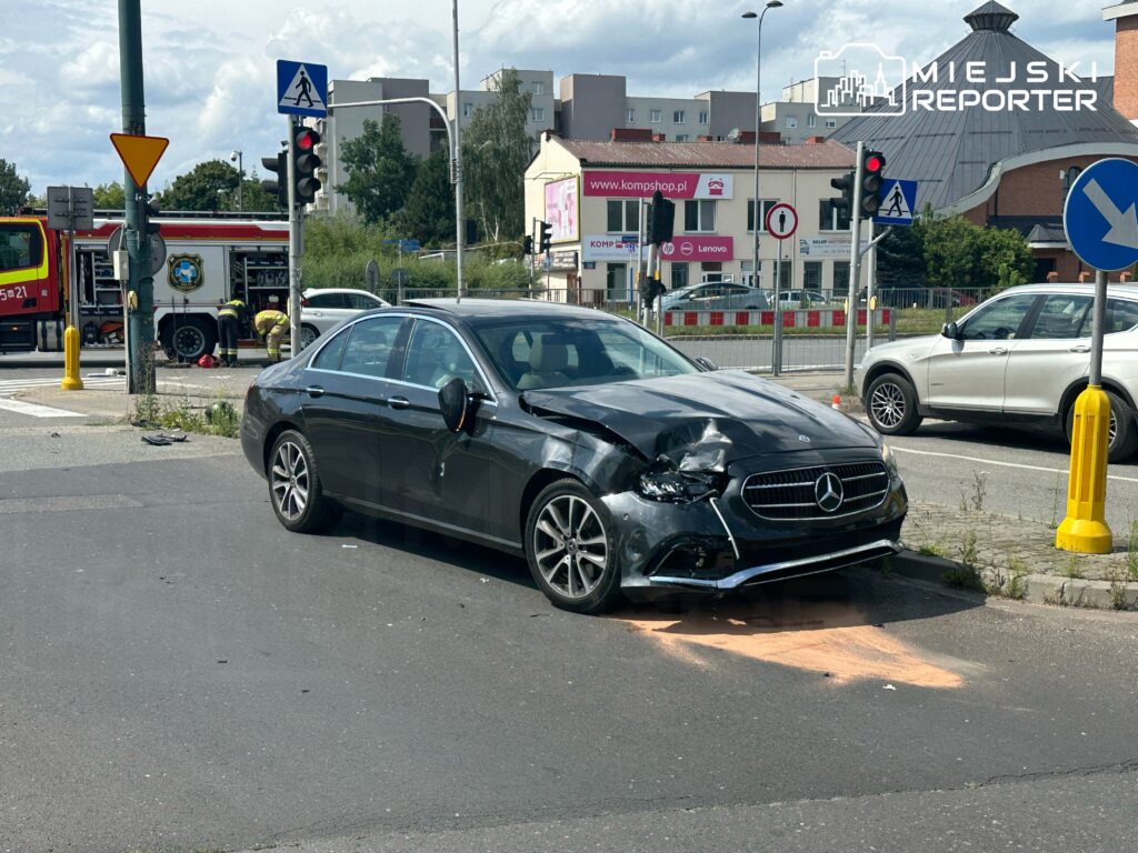 W zdarzeniu drogowym na skrzyżowaniu ulic doszło do kolizji dwóch samochodów. Na miejscu interweniują służby ratunkowe, w tym straż pożarna. Uszkodzony pojazd to czarny Mercedes z widocznymi zniszczeniami. Ruch w okolicy jest wstrzymany.