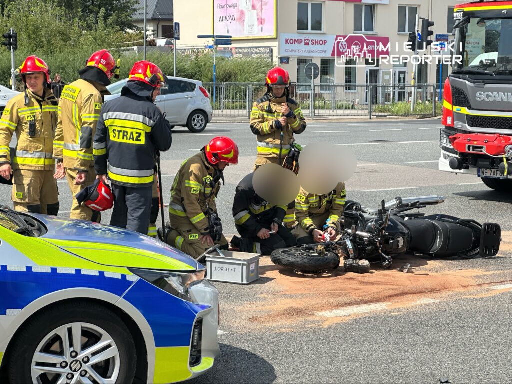 Na zdjęciu widać strażaków interweniujących na miejscu wypadku motocyklowego. Ratownicy udzielają pomocy poszkodowanemu, a obok leży przewrócony motocykl. W tle widoczny jest pojazd strażacki oraz ruch uliczny. Na miejscu zdarzenia widoczne są również oznaczenia służb ratunkowych.