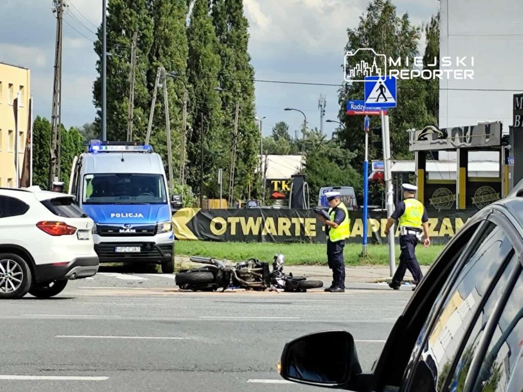 Na zdjęciu widać policyjny radiowóz oraz funkcjonariuszy zabezpieczających miejsce zdarzenia drogowego. Na jezdni leży przewrócony motocykl, a obok stoją świadkowie. W tle widoczna jest reklama oraz pieszy przejście. Zdarzenie miało miejsce w słoneczny dzień.