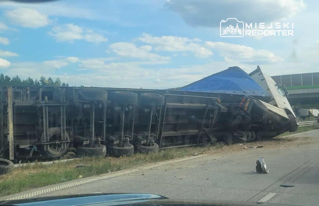 Na zdjęciu widoczny jest przewrócony ciężarówka na autostradzie. Pojazd leży na boku, a jego koła są do góry. W tle widać fragment jezdni oraz zieleń przydrożną. Sytuacja może powodować utrudnienia w ruchu drogowym. Na miejscu zdarzenia mogą być obecne służby ratunkowe.