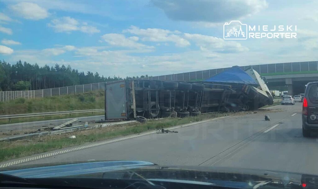 Na zdjęciu widać przewrócony ciężarówkę na autostradzie, obok której zablokowany jest ruch w kierunku przeciwnym. W tle znajdują się inne pojazdy oraz zieleń. Zdarzenie miało miejsce w słoneczny dzień, co może sugerować korzystne warunki pogodowe. Na miejscu są służby ratunkowe.