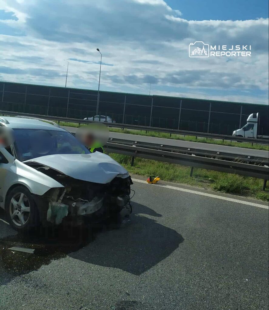 Wypadek drogowy na autostradzie. Zniszczony pojazd osobowy z uszkodzoną przednią częścią. Na miejscu zdarzenia obecni funkcjonariusze służb ratunkowych. W tle widoczne inne samochody i infrastruktura drogowa. Słoneczna pogoda. Informacje o przyczynach wypadku są w trakcie ustalania.