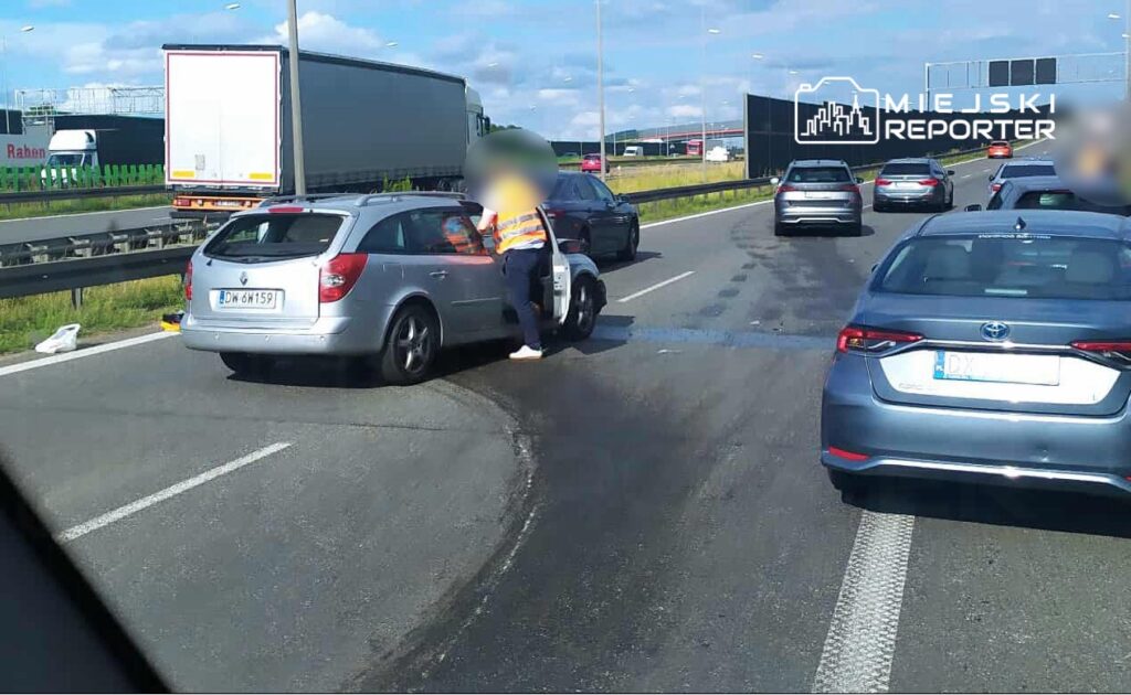Na drodze krajowej doszło do zdarzenia drogowego. Widać samochód osobowy zatrzymany na poboczu, obok niego stoi osoba w odblaskowej kamizelce. Ruch na jezdni jest spowolniony, a w tle widoczne są inne pojazdy. Na miejscu zdarzenia trwają czynności wyjaśniające.