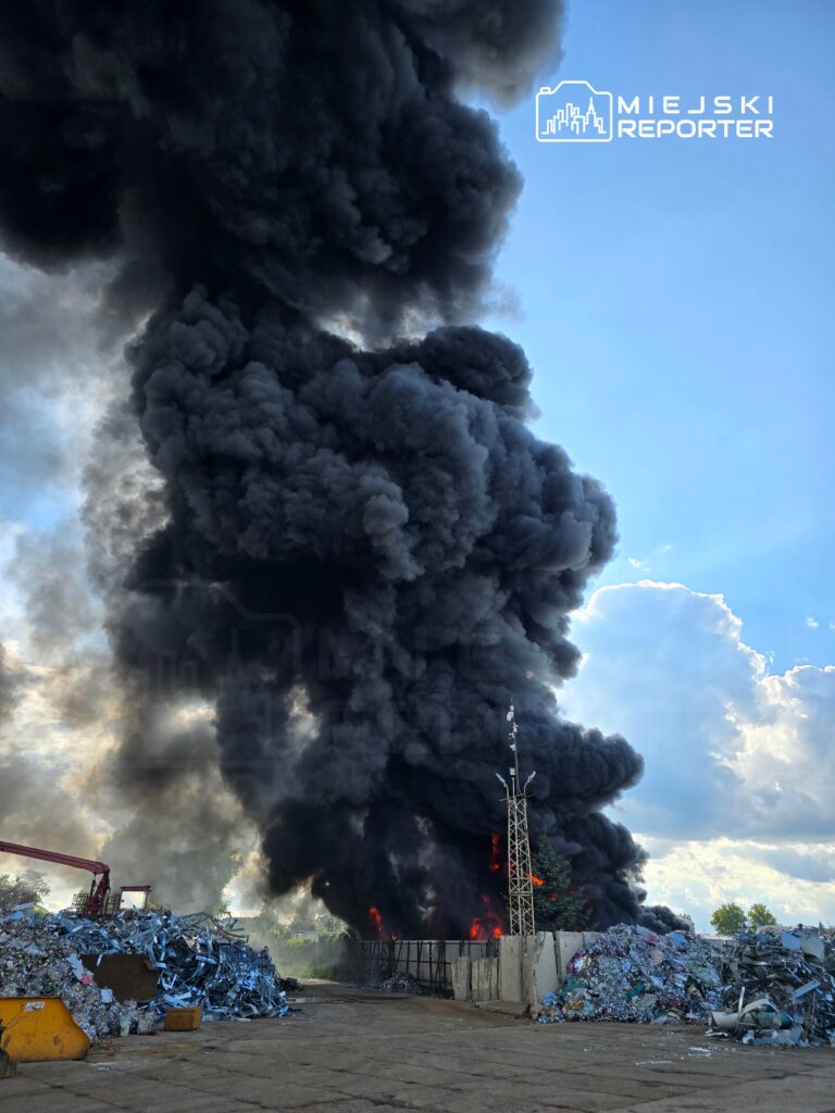 Wielka chmura czarnego dymu unosi się nad składowiskiem odpadów, gdzie doszło do pożaru. W tle widać płonące fragmenty oraz maszyny do przetwarzania odpadów. Miejsce zdarzenia jest otoczone metalowymi barierkami. Pożar wymaga interwencji służb ratunkowych.
