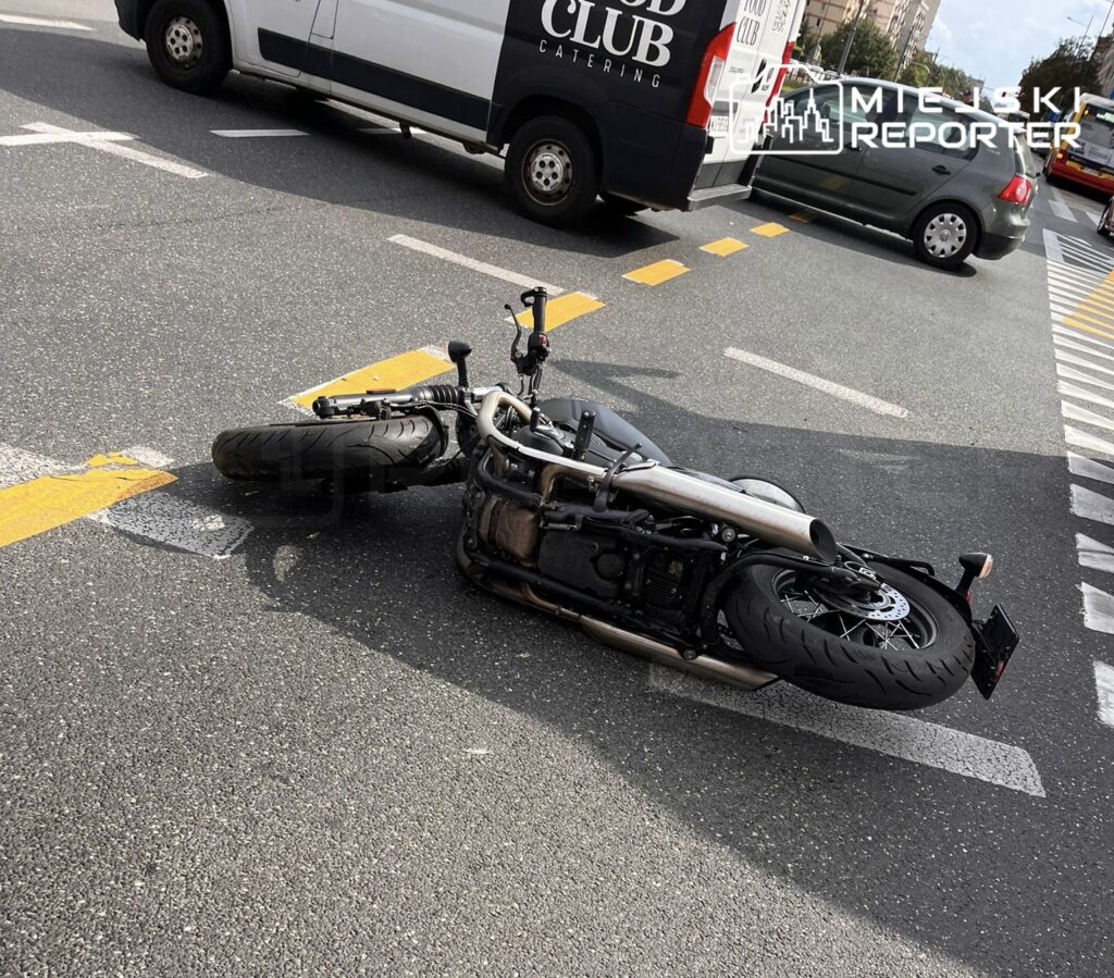 Motocykl leżący na jezdni, na skrzyżowaniu ulic. W tle widoczne pojazdy dostawcze oraz osobowe. Sytuacja sugeruje możliwy wypadek lub awarię jednośladu. Na drodze widoczne oznaczenia poziome. Zdjęcie dokumentuje zdarzenie drogowe w miejskim otoczeniu.
