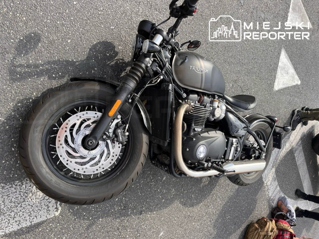 Motocykl marki Triumph leży na asfalcie, częściowo zasłonięty przez torbę i obuwie. W tle widoczni są ludzie, którzy mogą być świadkami zdarzenia. Obraz ilustruje sytuację po ewentualnym wypadku lub awarii jednośladu. Zdarzenie miało miejsce na drodze.