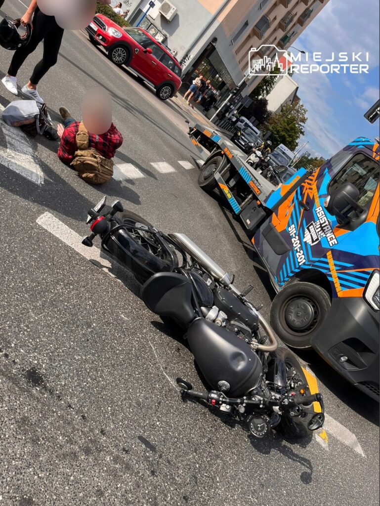 Wypadek drogowy z udziałem motocykla i osoby poszkodowanej. Motocykl leży na jezdni, a obok znajduje się ranny mężczyzna. Na miejscu zdarzenia obecni są świadkowie oraz służby ratunkowe. W tle widoczny pojazd pomocy drogowej oraz ruch samochodowy.