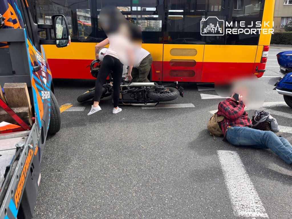 Wypadek drogowy z udziałem motocykla i autobusu miejskiego. Na miejscu zdarzenia osoby próbują pomóc poszkodowanemu motocykliście, który leży na jezdni. Widoczny również kierowca autobusu. W tle widać inne pojazdy. Służby ratunkowe mogą być w drodze.