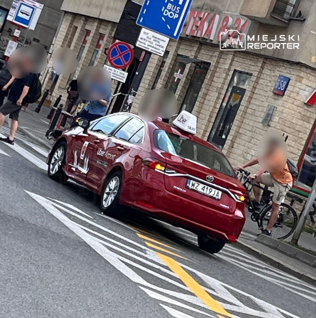Czerwony samochód osobowy z oznaczeniem taksówki Uber stoi na skrzyżowaniu w mieście. W tle widoczni są piesi oraz rowerzysta, a także znaki drogowe informujące o przystanku autobusowym i zakazie parkowania. Scena przedstawia ruch uliczny w miejskim otoczeniu.