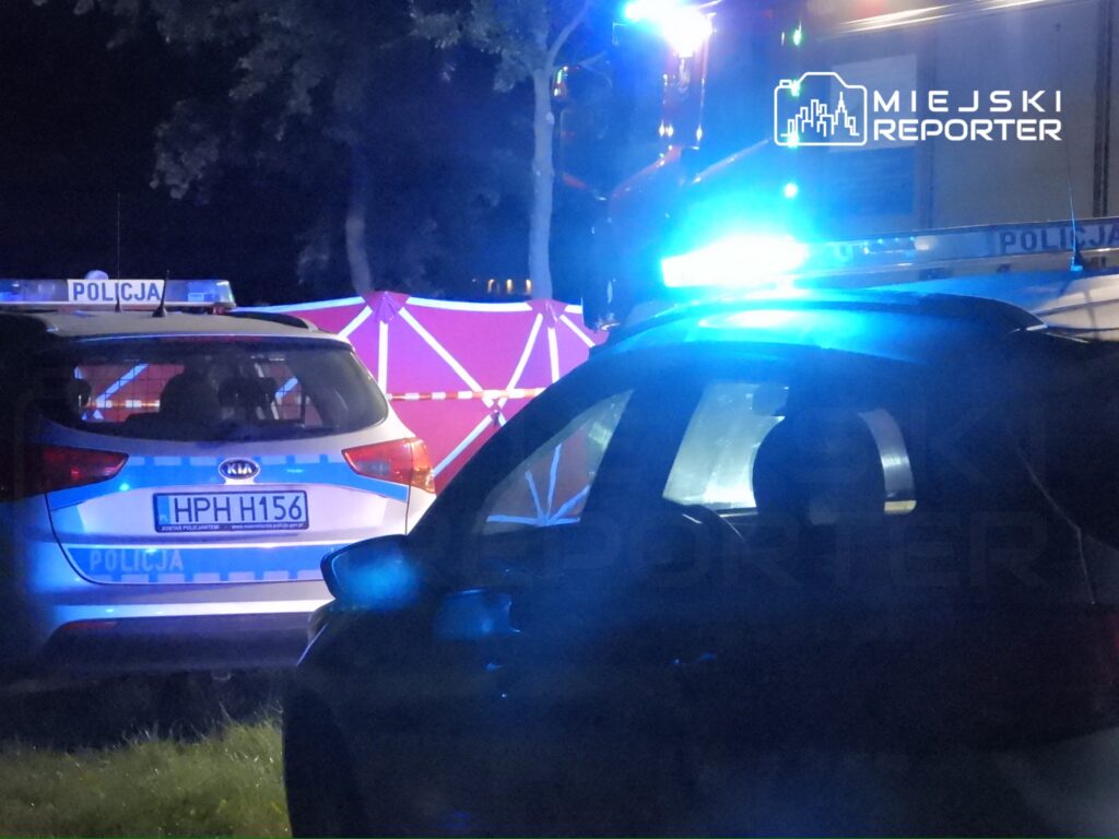 Na zdjęciu widoczna jest akcja policyjna w nocy. Dwa radiowozy z włączonymi sygnałami świetlnymi oraz zabezpieczony teren, co wskazuje na prowadzenie dochodzenia w danym miejscu. Obecność służb porządkowych sugeruje, że miało miejsce zdarzenie wymagające interwencji.