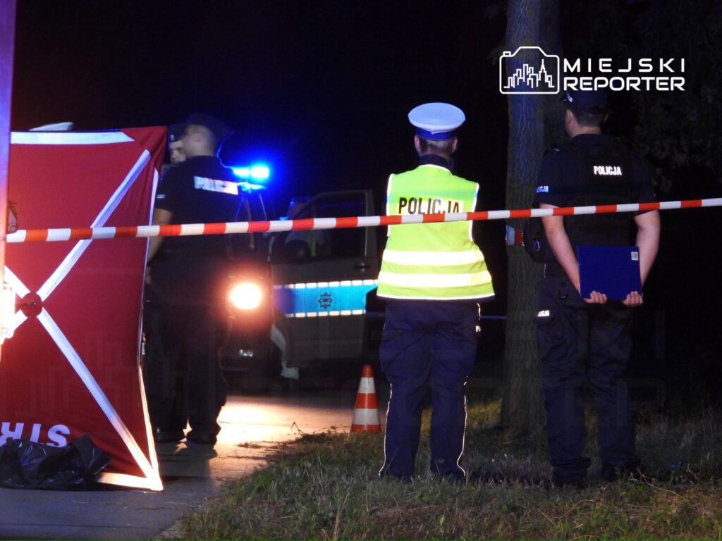 Policjanci zabezpieczają miejsce zdarzenia w nocy. Na pierwszym planie widoczny jest funkcjonariusz w kamizelce odblaskowej oraz dwóch policjantów, którzy rozmawiają. W tle radiowóz policyjny z włączonymi sygnałami świetlnymi. Obszar otoczony taśmą zabezpieczającą.