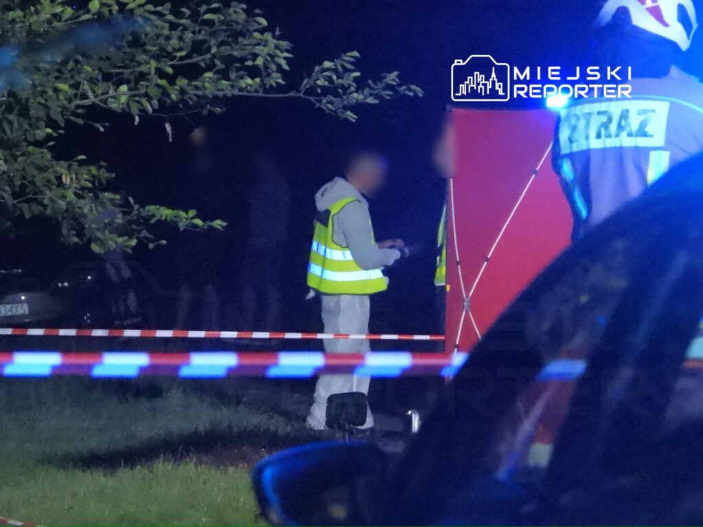 Na zdjęciu widać pracowników służb ratunkowych w kamizelkach odblaskowych, którzy prowadzą działania na miejscu zdarzenia w nocy. W tle widoczne są oznaczenia policyjne oraz zablokowany obszar. Sytuacja wskazuje na interwencję w związku z incydentem wymagającym wsparcia służb.