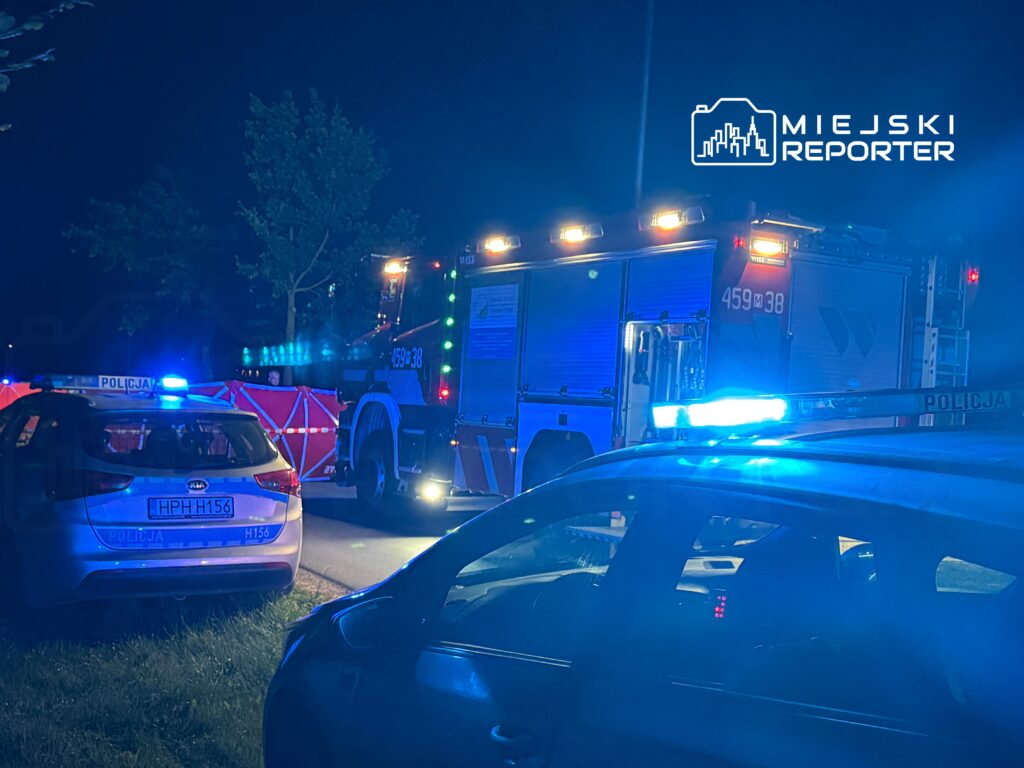 Na zdjęciu widoczny jest interwencyjny pojazd strażacki oraz radiowozy policyjne, oświetlone niebieskimi sygnalizacjami. Scena rozgrywa się w nocy, w tle znajdują się drzewa. Obraz dokumentuje działania służb ratunkowych w trakcie zdarzenia.