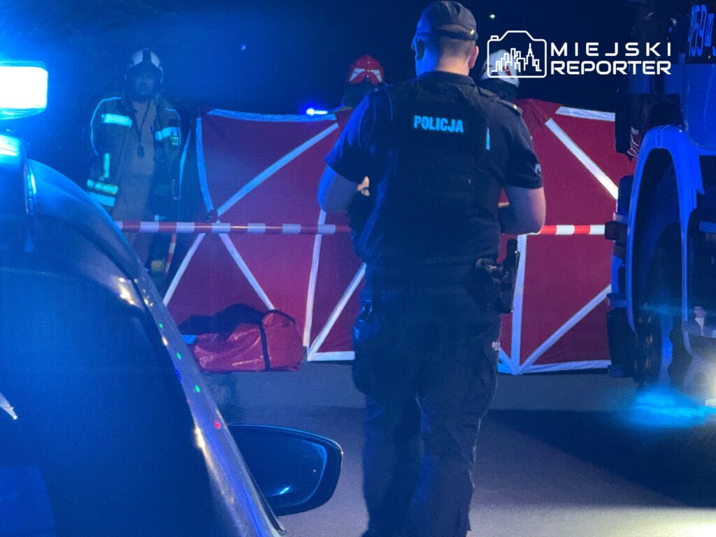 Na zdjęciu widać interwencję służb ratunkowych i policji w miejscu zdarzenia. Funkcjonariusze zabezpieczają teren, a w tle widoczna jest czerwona bariera. W akcji biorą udział strażacy oraz policjanci, co sugeruje poważny incydent. W nocy, przy niebieskim świetle radiowozów, sytuacja budzi niepokój.