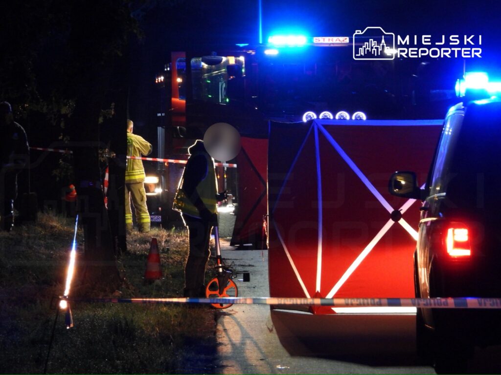 Na zdjęciu przedstawiono akcję ratunkową prowadzoną przez strażaków i policję w nocy. W tle widoczny jest wóz strażacki oraz oznakowane miejsce zdarzenia. Funkcjonariusze zabezpieczają teren i prowadzą działania związane z incydentem drogowym. Widać również sprzęt pomiarowy.