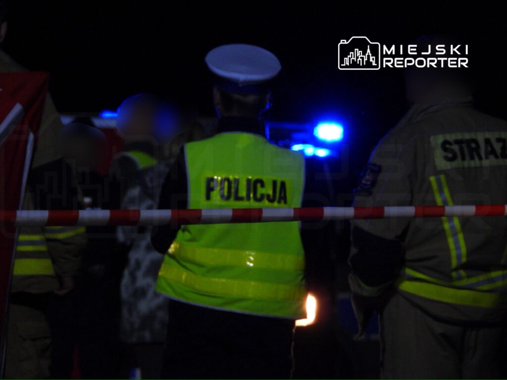 Na zdjęciu widoczna jest interwencja służb mundurowych w nocy. Funkcjonariusze policji oraz straży pożarnej prowadzą działania na miejscu zdarzenia, zabezpieczając teren. W tle migają niebieskie światła radiowozu. Scena wskazuje na poważny incydent wymagający obecności służb ratunkowych.