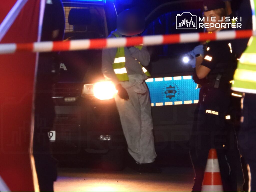 Na zdjęciu widać funkcjonariuszy policji oraz pracowników służb ratunkowych na miejscu zdarzenia. W tle znajduje się radiowóz z włączonymi sygnałami świetlnymi. Obszar jest zabezpieczony taśmą, a w pobliżu znajdują się pachołki drogowe. Akcja ma miejsce w nocy.