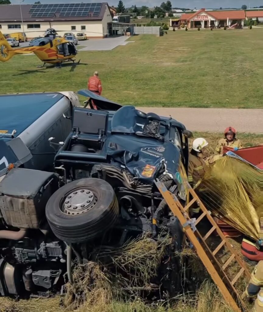 Pożarnicy pracują przy przewróconym ciężarówce, a helikopter lądował w tle na trawiastej przestrzeni.