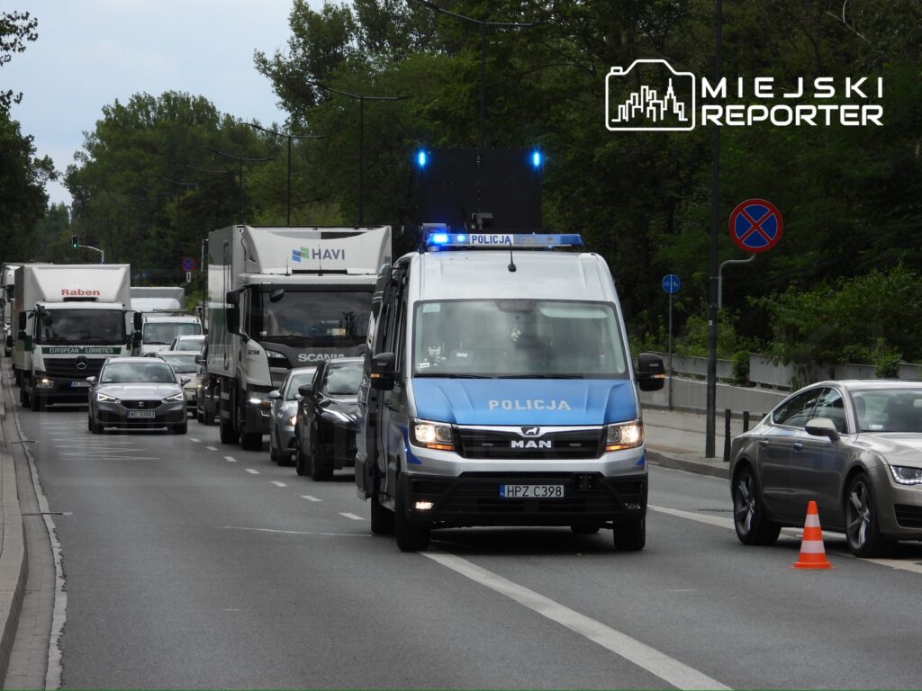 Policyjny pojazd z niebieskim światłem sygnalizacyjnym przejeżdża przez zator drogowy w miejskim otoczeniu.