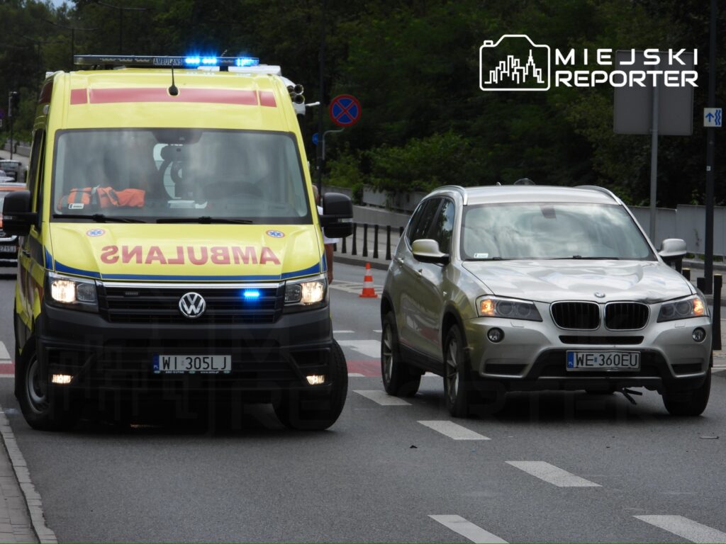 Ambulans z niebieskimi światłami przejeżdża obok zaparkowanego samochodu osobowego na ulicy.